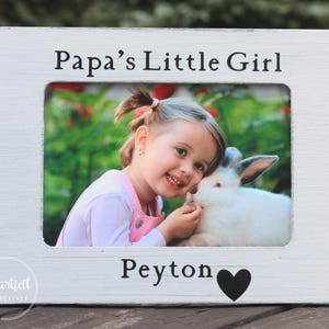 May include: White wooden picture frame with a photo of a young girl with a rabbit. The frame has the text "Papa's Little Girl" at the top and "Peyton" with a heart at the bottom.