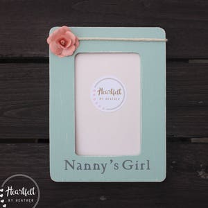Puede incluir: Un marco de fotos de madera azul claro con un adorno de flor rosa. El marco tiene un colgador de cuerda blanca y el texto "Nanny's Girl" está impreso en la parte inferior. El marco tiene una pegatina con el texto "Heartfelt by Heather".