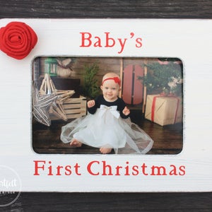 Puede incluir: Marco de fotos de madera blanca con una rosa de tela roja en la esquina superior izquierda. El marco tiene las palabras "Baby's First Christmas" en letras rojas. El marco contiene una foto de una niña en un vestido blanco sentada frente a un árbol de Navidad.