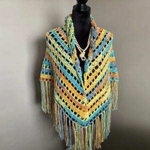 May include: A crocheted shawl with a colorful striped pattern and fringe. The shawl is displayed on a mannequin with a pearl necklace.