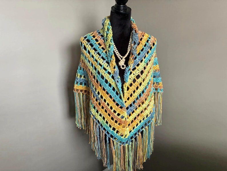May include: A crocheted shawl with a colorful striped pattern and fringe. The shawl is displayed on a mannequin with a pearl necklace.