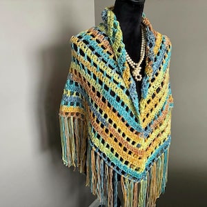 May include: A crocheted shawl with a fringe border in shades of blue, green, yellow, and orange. The shawl is displayed on a mannequin.