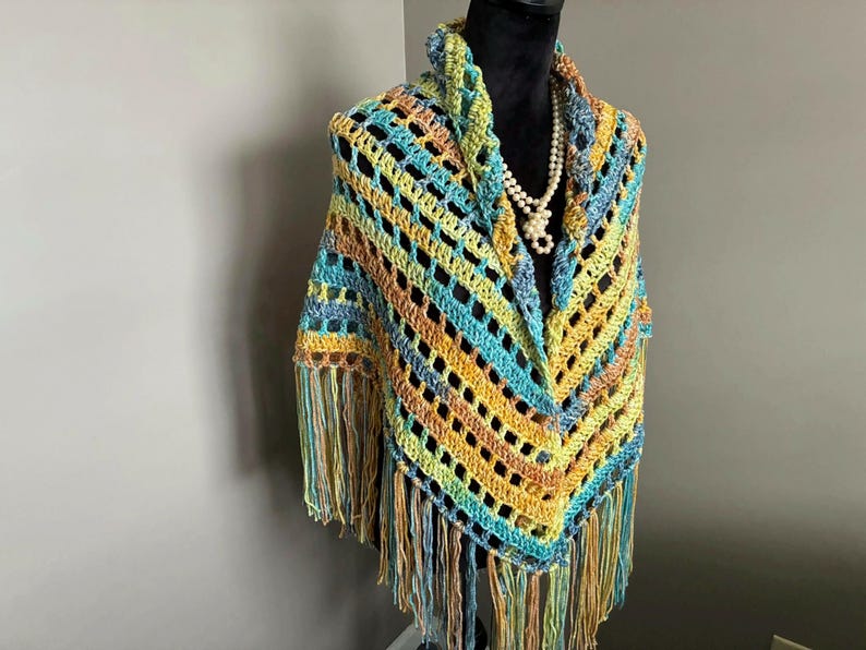 May include: A crocheted shawl with a fringe border in shades of blue, green, yellow, and orange. The shawl is displayed on a mannequin.