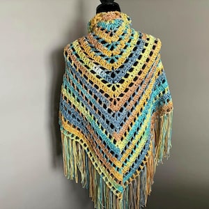 May include: A crocheted shawl with a colorful striped pattern in shades of blue, green, yellow, and orange. The shawl has a fringe border and is displayed on a mannequin.
