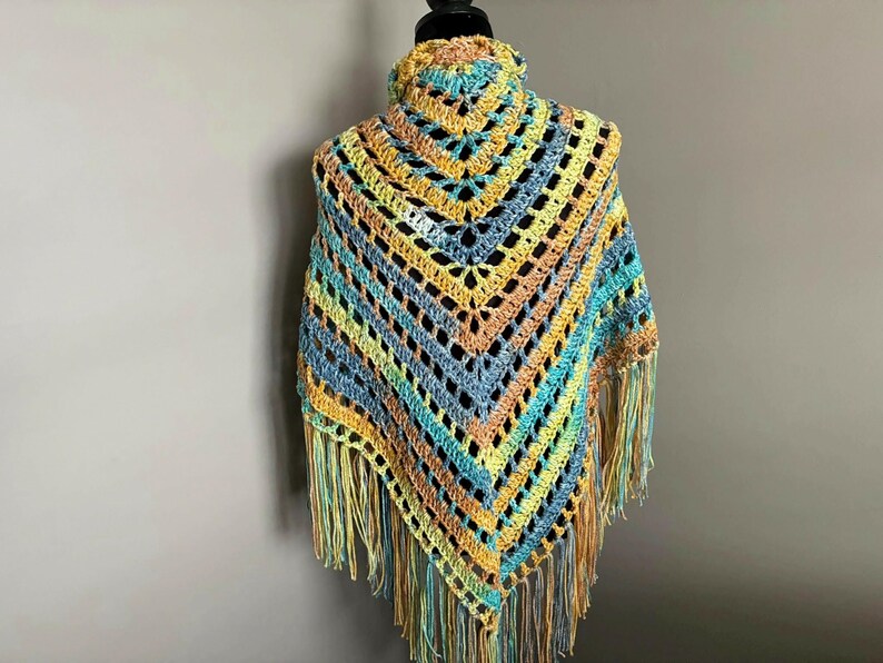 May include: A crocheted shawl with a colorful striped pattern in shades of blue, green, yellow, and orange. The shawl has a fringe border and is displayed on a mannequin.