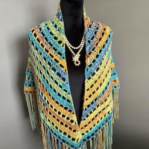 May include: A crocheted shawl with a fringe border in shades of teal, yellow, and brown. The shawl is draped over a mannequin wearing a pearl necklace.