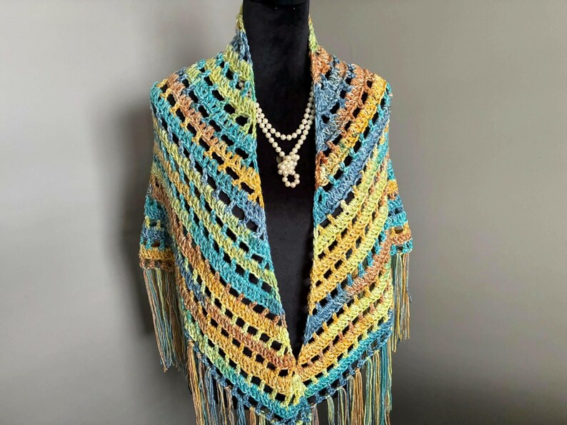 May include: A crocheted shawl with a fringe border in shades of teal, yellow, and brown. The shawl is draped over a mannequin wearing a pearl necklace.