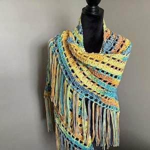 May include: A crocheted shawl with a fringe border in shades of blue, green, and yellow. The shawl is draped over a mannequin.