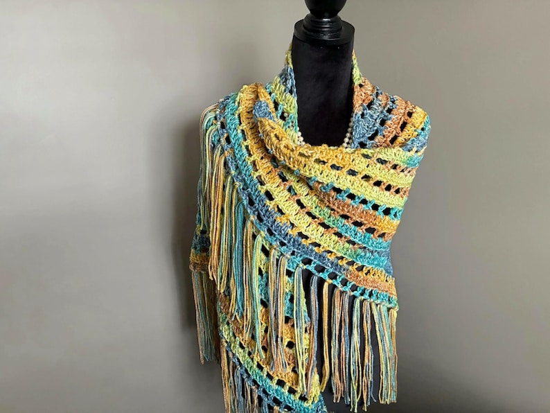 May include: A crocheted shawl with a fringe border in shades of blue, green, and yellow. The shawl is draped over a mannequin.