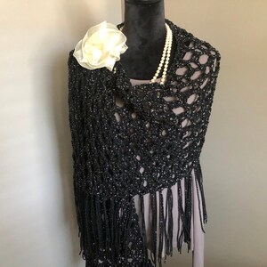 May include: A black crochet shawl with fringe, featuring a delicate openwork pattern. A white flower brooch is pinned to the shawl.