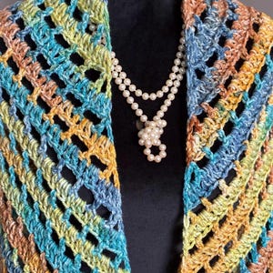 May include: A crocheted shawl in shades of blue, green, and orange with a fringe border. The shawl is draped over a mannequin wearing a pearl necklace.