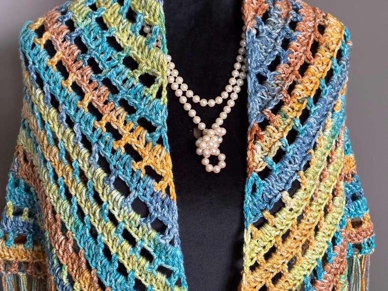 May include: A crocheted shawl in shades of blue, green, and orange with a fringe border. The shawl is draped over a mannequin wearing a pearl necklace.