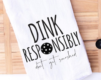 Pode incluir: Toalha de cozinha branca com texto preto que diz "Dink Responsibly don't get smashed" com uma imagem em preto e branco de uma bola de pickleball no meio do texto.
