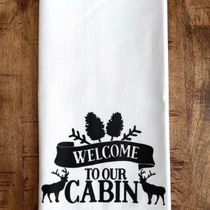May include: White kitchen towel with black text and images. The text reads "Welcome to our Cabin" with a banner and two deer silhouettes.