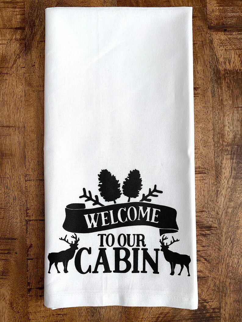 May include: White kitchen towel with black text and images. The text reads "Welcome to our Cabin" with a banner and two deer silhouettes.