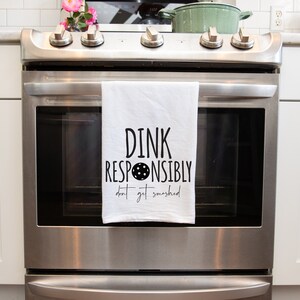 May include: A white kitchen towel with black text that reads "DINK RESPONSIBLY don't get smashed" with a black circle and dots in the middle of the word "RESPONSIBLY". The towel is hanging on a stainless steel oven.
