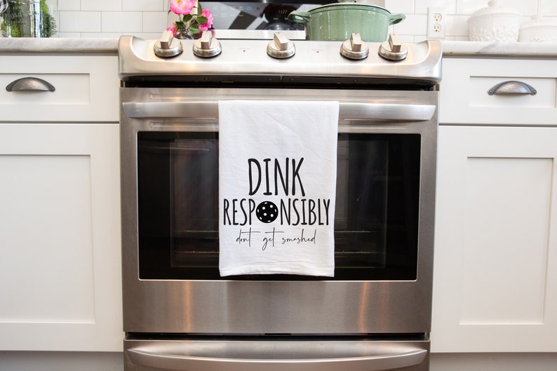 May include: A white kitchen towel with black text that reads "DINK RESPONSIBLY don't get smashed" with a black circle and dots in the middle of the word "RESPONSIBLY". The towel is hanging on a stainless steel oven.