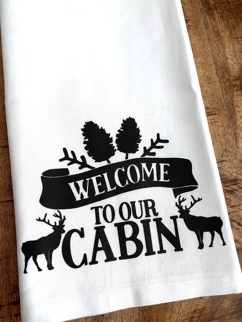 May include: White kitchen towel with black text and images. The text reads "Welcome to our Cabin". There are two pine trees and two elk silhouettes.