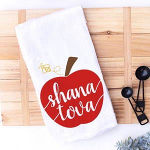 May include: White kitchen towel featuring a red apple design. The apple has the text "shana tova" written inside. A small yellow bee is flying above the apple.