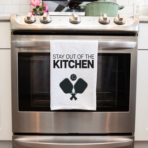 May include: A white kitchen towel with black text that reads "Stay out of the kitchen" and a black silhouette of two pickleball paddles and a ball.