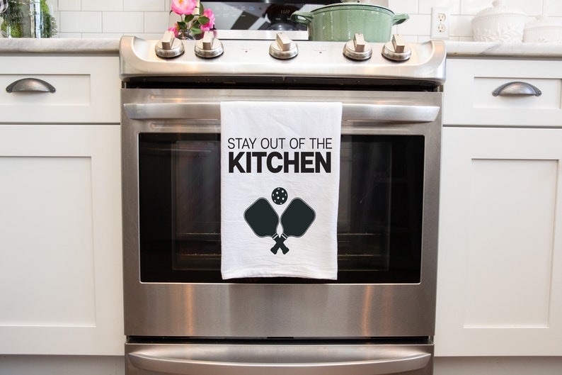May include: A white kitchen towel with black text that reads "Stay out of the kitchen" and a black silhouette of two pickleball paddles and a ball.