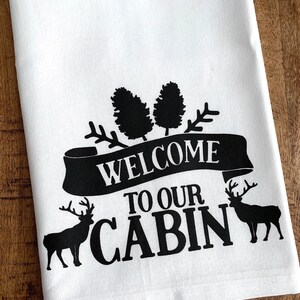 May include: White kitchen towel with black text that reads "Welcome to our Cabin". The text is surrounded by pine trees and two deer silhouettes.