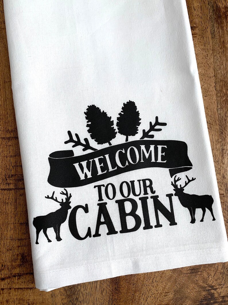 May include: White kitchen towel with black text that reads "Welcome to our Cabin". The text is surrounded by pine trees and two deer silhouettes.