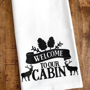 May include: White kitchen towel with black text and images. The text reads "Welcome to our Cabin". There are two pine trees and two deer silhouettes.