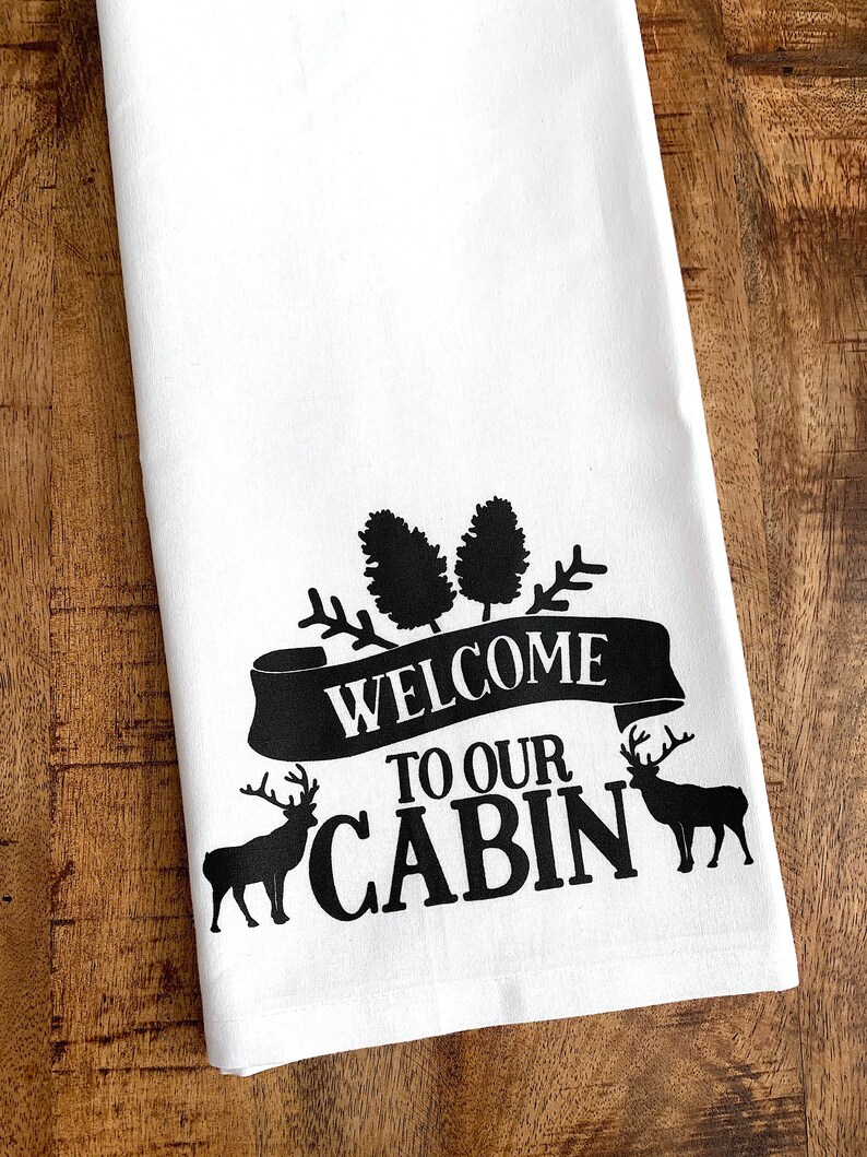 May include: White kitchen towel with black text and images. The text reads "Welcome to our Cabin". There are two pine trees and two deer silhouettes.