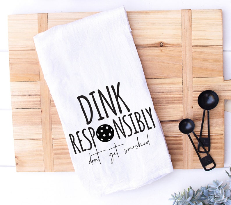 May include: White kitchen towel with black text that reads "Dink Responsibly don't get smashed" with a black and white image of a pickleball in the middle of the text.