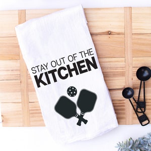 May include: White kitchen towel with black text that reads "Stay out of the kitchen" and a black silhouette of two pickleball paddles and a ball.