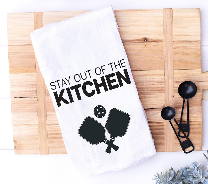 May include: White kitchen towel with black text that reads "Stay out of the kitchen" and a black silhouette of two pickleball paddles and a ball.