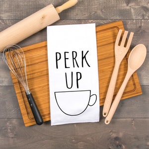 May include: White tea towel with the text "PERK UP" and a black outline of a coffee cup. The tea towel is on a wooden cutting board with a whisk, rolling pin, and wooden spoons.