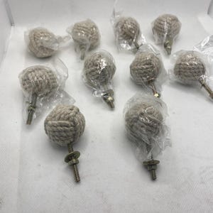 May include: A collection of ten beige rope knot cabinet knobs. Each knob is wrapped in clear plastic and has a threaded metal screw for installation. The knobs are designed for furniture and home decor.