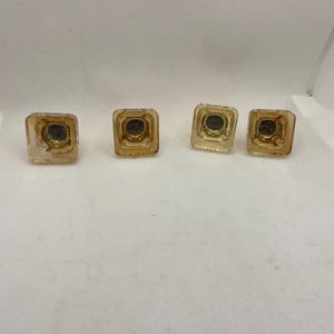 May include: Four square, amber-colored glass cabinet knobs. Each knob has a dark, circular center and a slightly rounded top. The knobs are evenly spaced against a white background, suggesting a hardware or home decor theme.