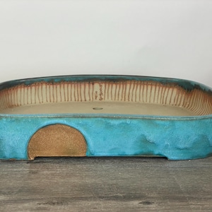 Puede incluir: Una jardinera rectangular de cerámica con esquinas redondeadas. El exterior es de un turquesa vibrante, mientras que el interior tiene una base beige con líneas marrones verticales. Se ve un recorte semicircular en un lado.