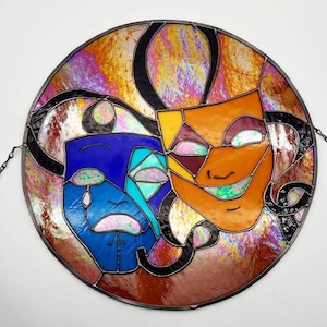 May include: A round stained glass piece featuring the classic comedy and tragedy masks. The masks are orange and blue, with black outlines and a colorful, iridescent background. A chain is attached for hanging.