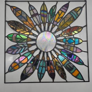 May include: A square stained glass panel featuring a central iridescent circle surrounded by colorful, feather-shaped elements. The feathers display hues of orange, yellow, green, blue, and purple, with a black metal frame.