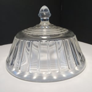 May include: Clear glass cloche with a ribbed, flared base and a domed lid. The lid has a small, decorative finial. The glass is transparent, allowing light to pass through. This decorative piece is suitable for displaying small items.