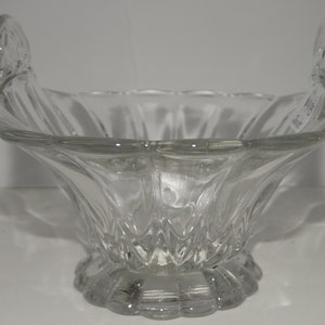 May include: Clear glass serving bowl with two curved handles. The bowl has a scalloped base and a flared, textured design. The handles are also textured and curve upwards. The bowl is designed to hold food items.