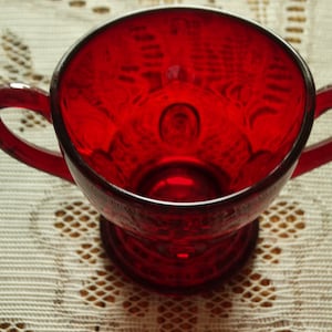 May include: A ruby red glass sugar bowl with two handles. The bowl has a pedestal base and a detailed pattern on the exterior. The sugar bowl is empty and sits on a white lace tablecloth.