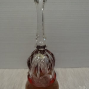 May include: A clear glass bell with a long, faceted handle and a bell-shaped body. The bell has a reddish-brown hue and is decorated with cut glass patterns. The bell is likely a decorative item.