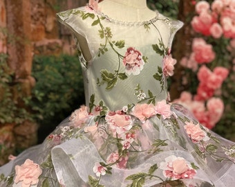 May include: A white tulle dress with a green floral print and pink 3D flowers. The dress has a sheer bodice and a full skirt.