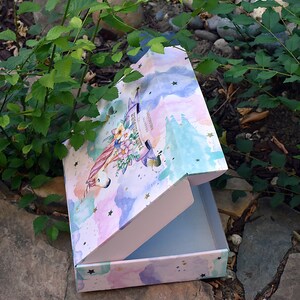 Pu&ograve; includere: Una scatola regalo rosa e blu con un design di unicorno e fiori. La scatola &egrave; aperta e ha un interno bianco. La scatola ha stelle dorate e il testo "La pi&ugrave; grande avventura &egrave; essere te stesso".
