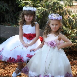 May include: Two young girls wearing white dresses with flower embellishments. The girl on the left is wearing a red sash and a white flower crown. The girl on the right is wearing a pink sash and a purple flower crown.