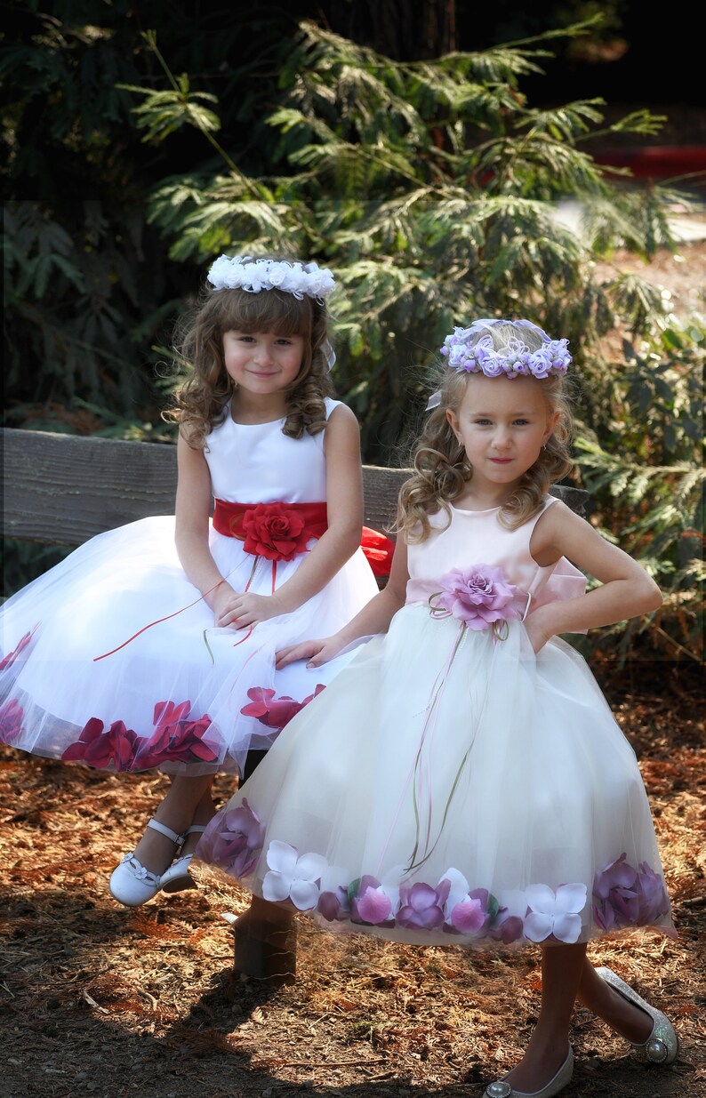 May include: Two young girls wearing white dresses with flower embellishments. The girl on the left is wearing a red sash and a white flower crown. The girl on the right is wearing a pink sash and a purple flower crown.