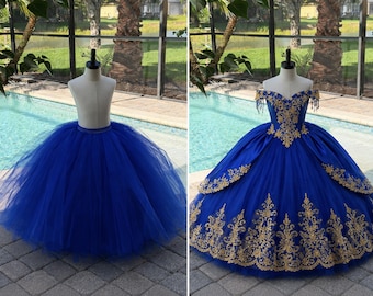 May include: A royal blue tulle skirt and a matching ball gown. The ball gown features gold embroidery on the bodice and skirt. The dresses are displayed on mannequins, set against a backdrop of a pool and palm trees.
