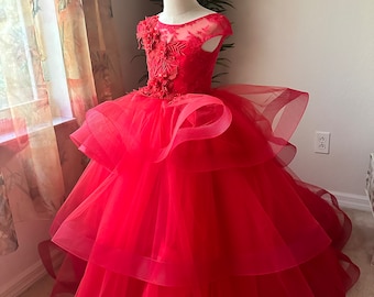 May include: A vibrant red formal dress with a layered skirt and floral bodice. The dress features a sheer neckline and is displayed on a mannequin. The skirt has multiple tiers of flowing fabric, creating a full, elegant silhouette. The dress is designed for special occasions.