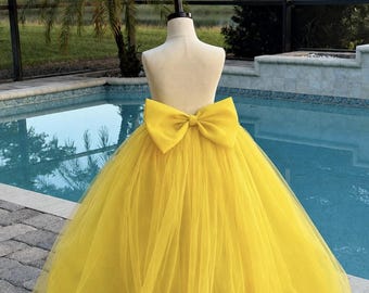 May include: A bright yellow floor-length skirt featuring multiple layers of sheer tulle fabric. The skirt is designed with a high waist and includes a large, prominent matching yellow bow attached at the center back of the waistband.