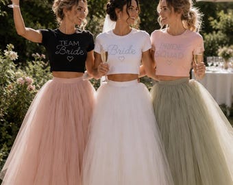 May include: Three cropped t-shirts paired with voluminous tulle midi skirts. The black shirt reads "TEAM Bride" with a heart, the white shirt reads "Bride" with a heart, and the light pink shirt reads "BRIDE SQUAD" with a heart.